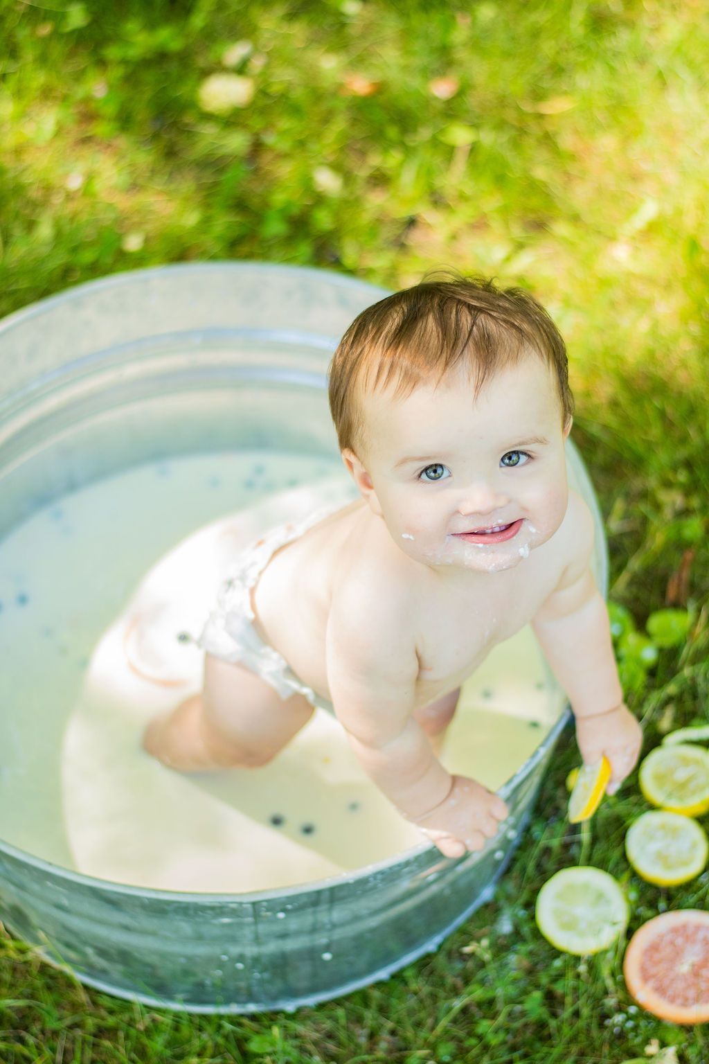 Baby smiling in a metal tub outdoors with water and sliced citrus nearby