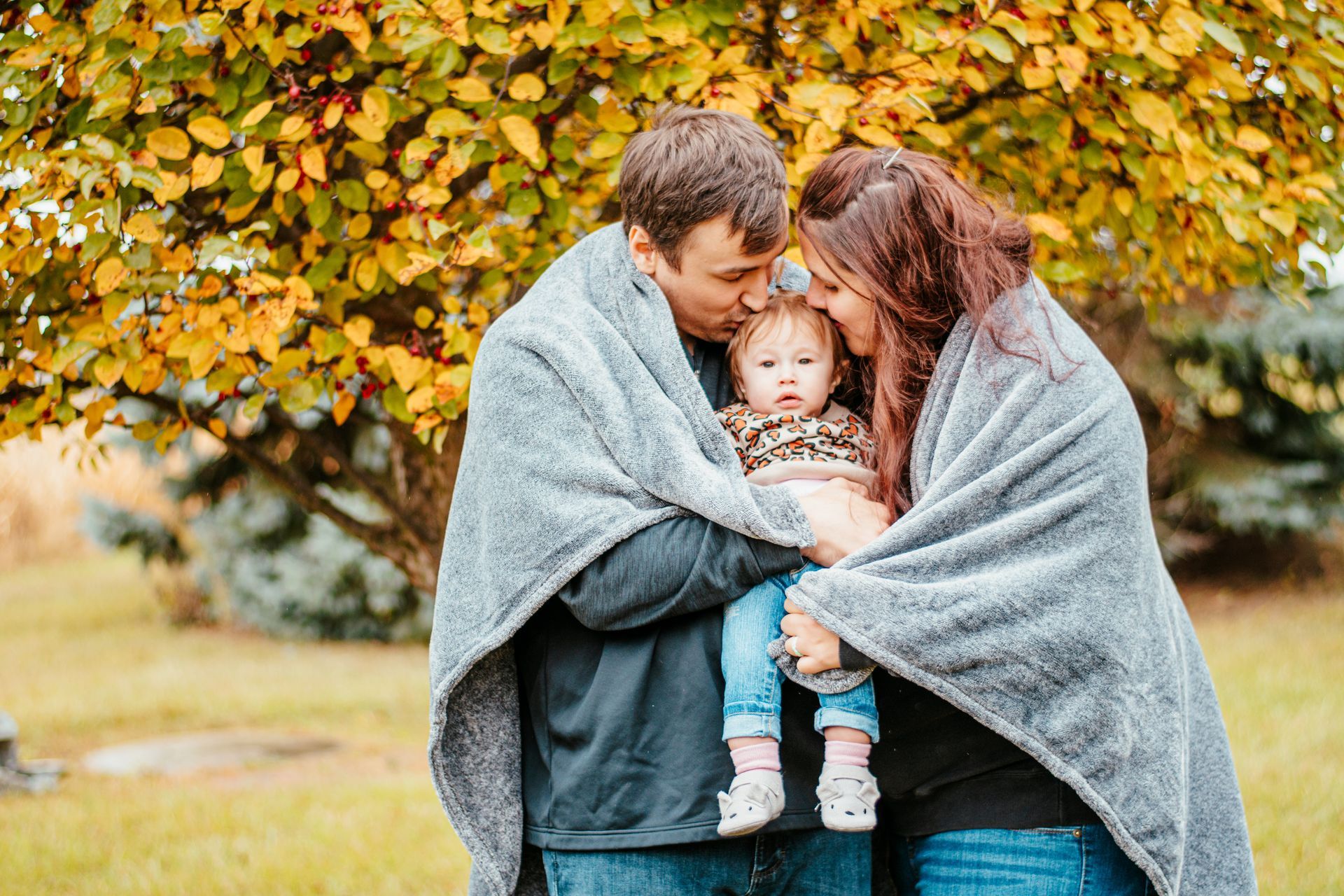 Two adults wrapped in gray blankets holding a baby outdoors under autumn leaves