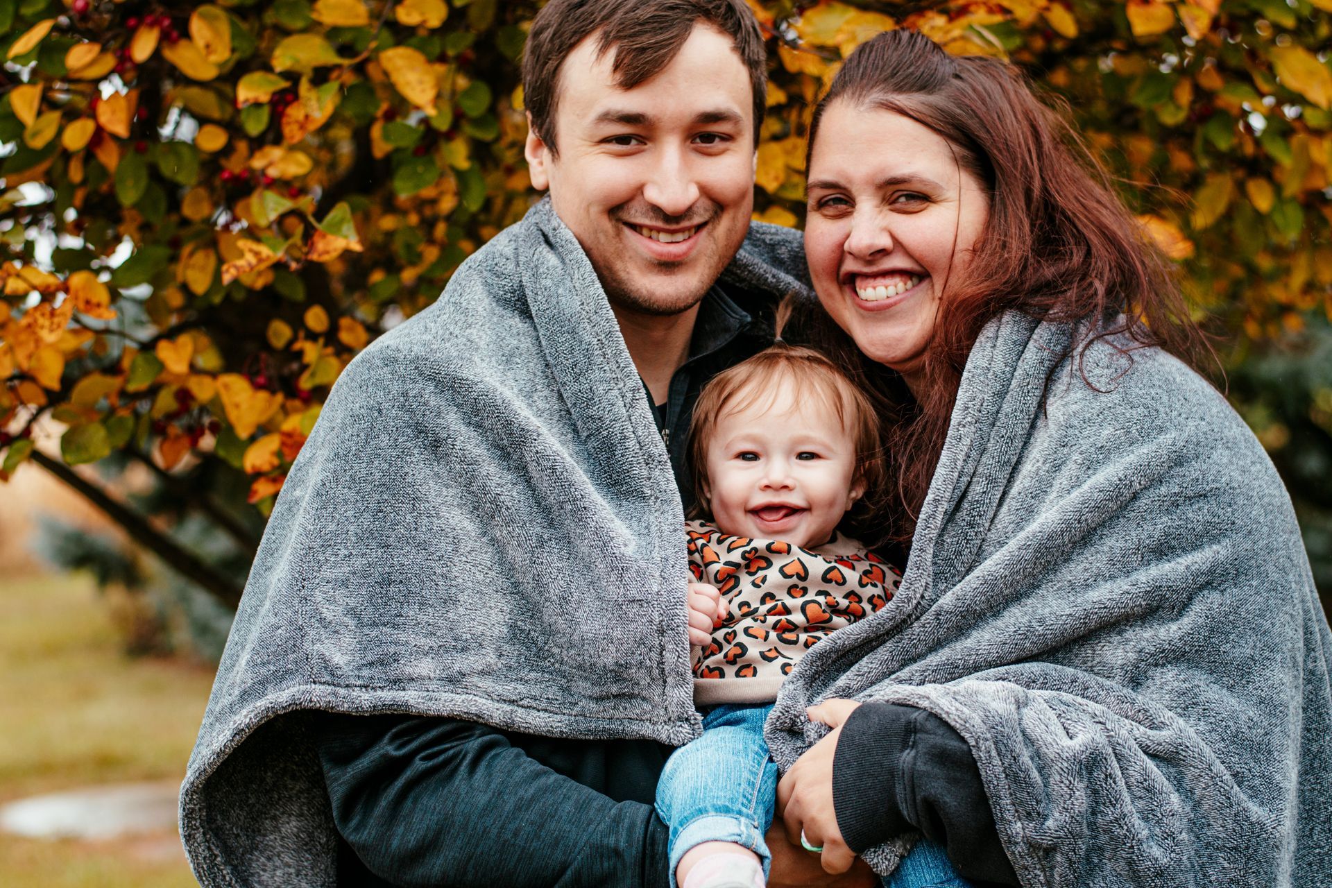 Smiling family of three wrapped in a gray blanket outdoors with autumn leaves behind them