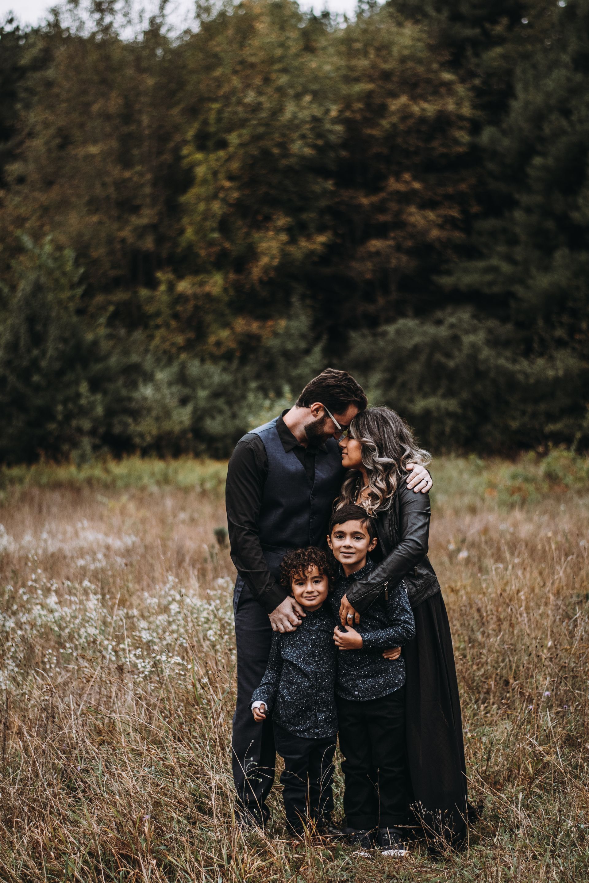Family of four embracing in a grassy field with trees in the background, dressed in dark clothing.