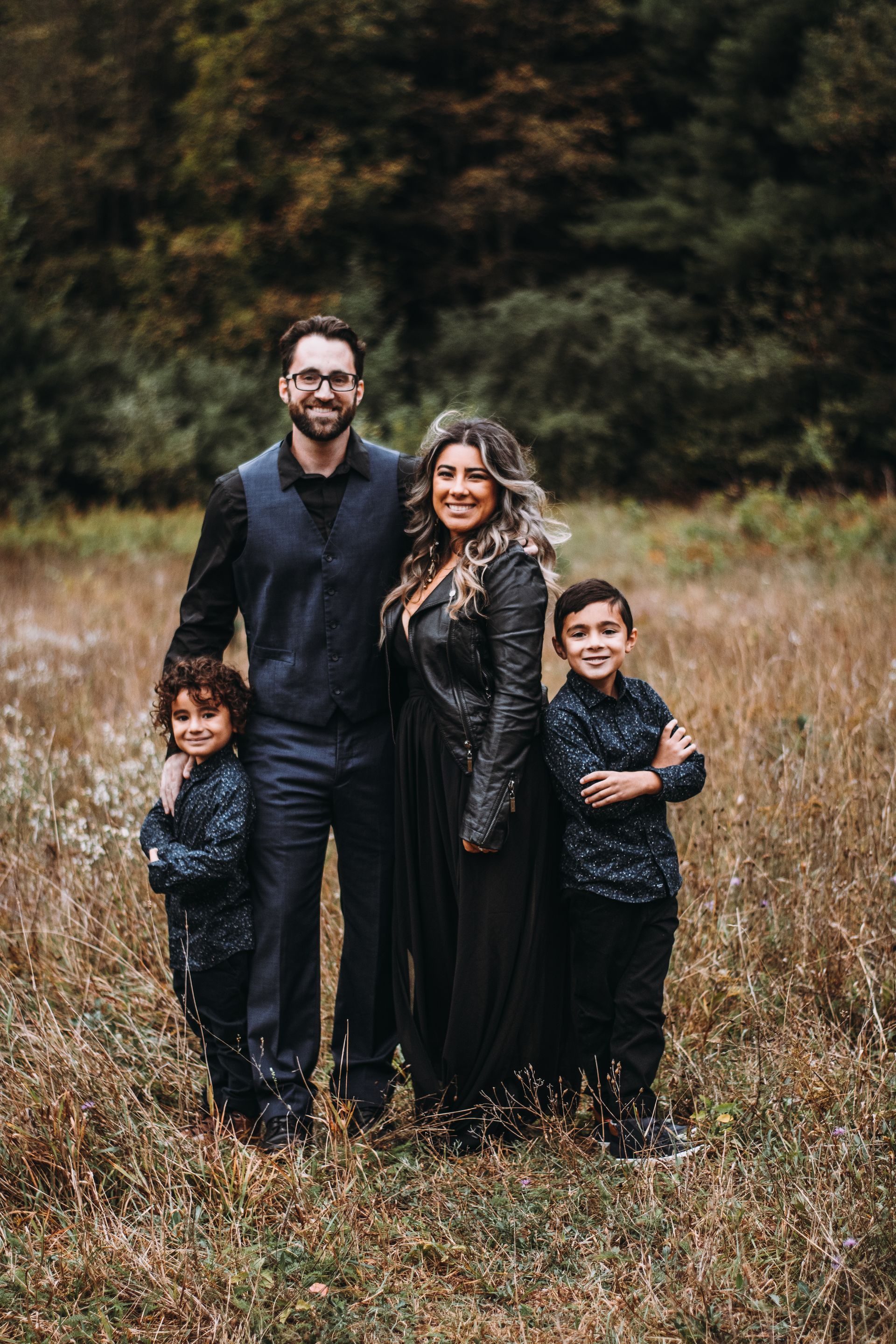 Family of four standing in a grassy field, wearing dark clothing, with a blurred forest backdrop.