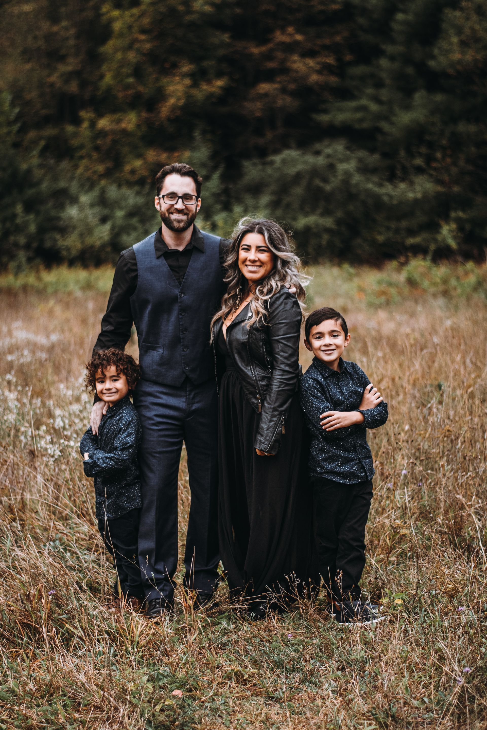 Family of four standing in a grassy field, wearing dark outfits, with a forest backdrop.