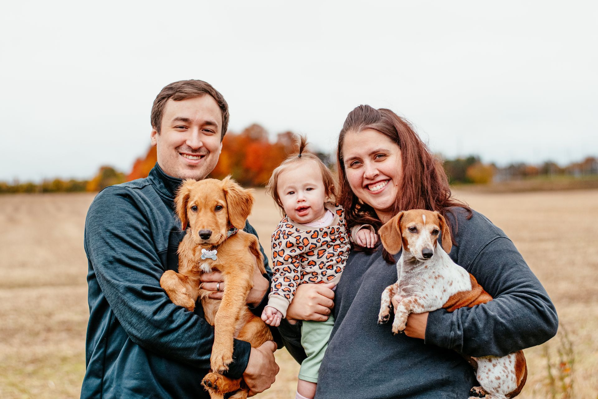 Family smiling outdoors in a field, holding two dogs and a baby