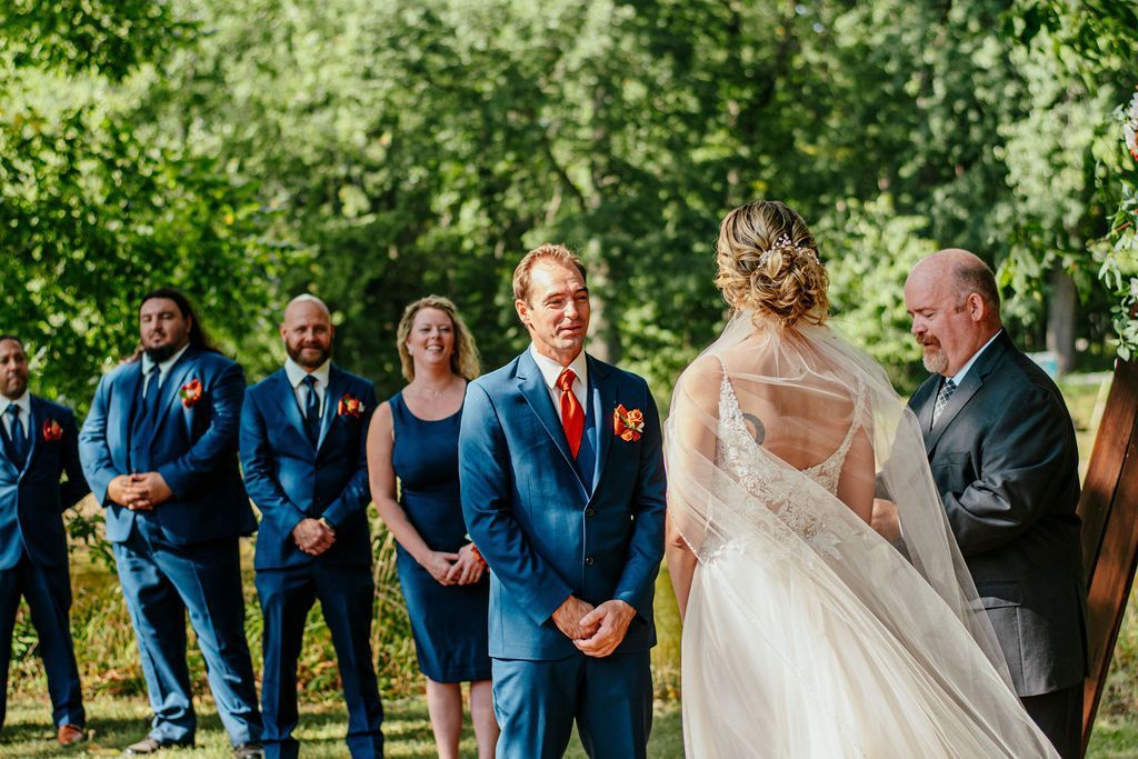 Wedding party outdoors in blue suits, facing the bride in a white veil and dress among trees.