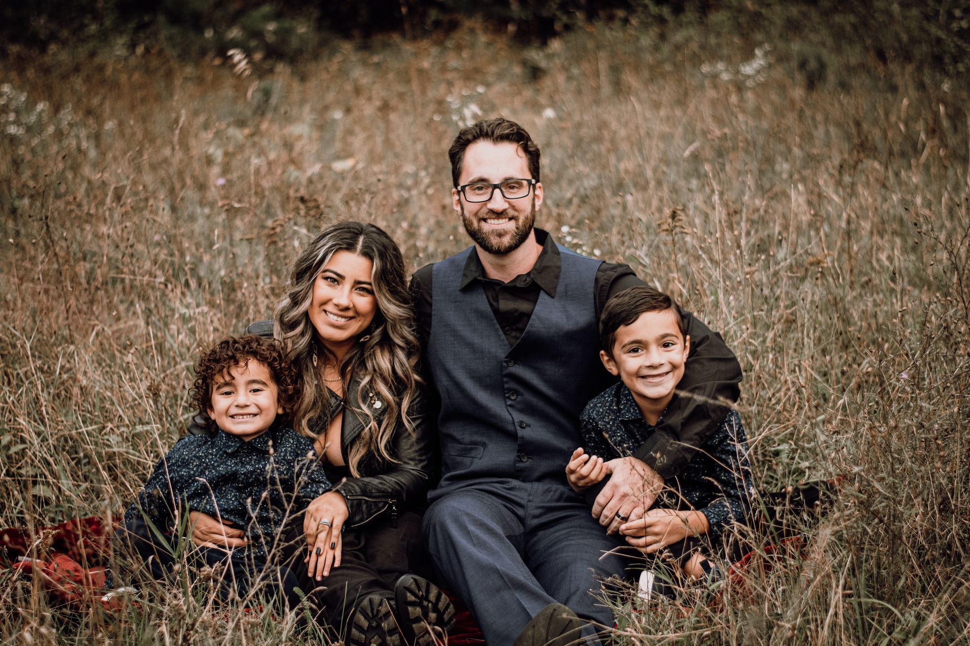 Family of four sitting together in tall grass, smiling outdoors.