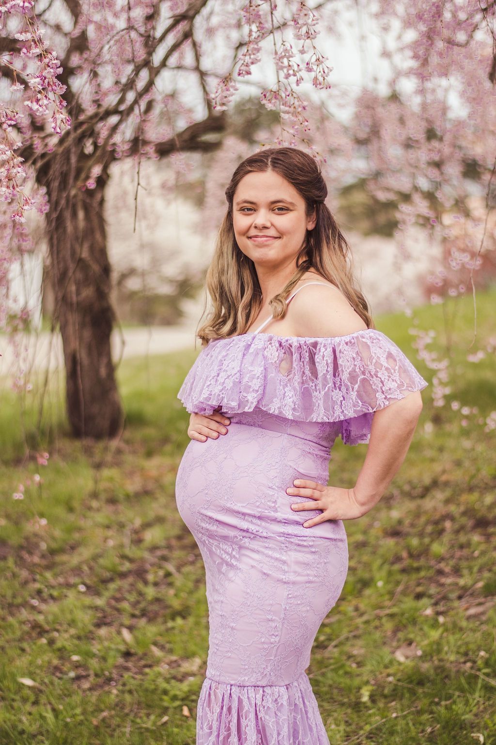 Pregnant woman in a lavender dress smiling outdoors among blooming trees