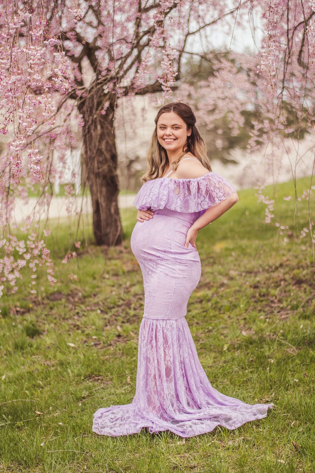 Pregnant woman in a lavender dress posing outdoors under pink cherry blossoms