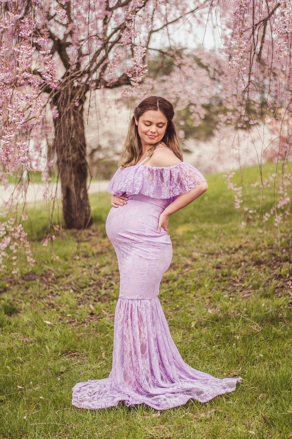 Pregnant woman in a lavender dress standing under blooming pink trees in a grassy orchard