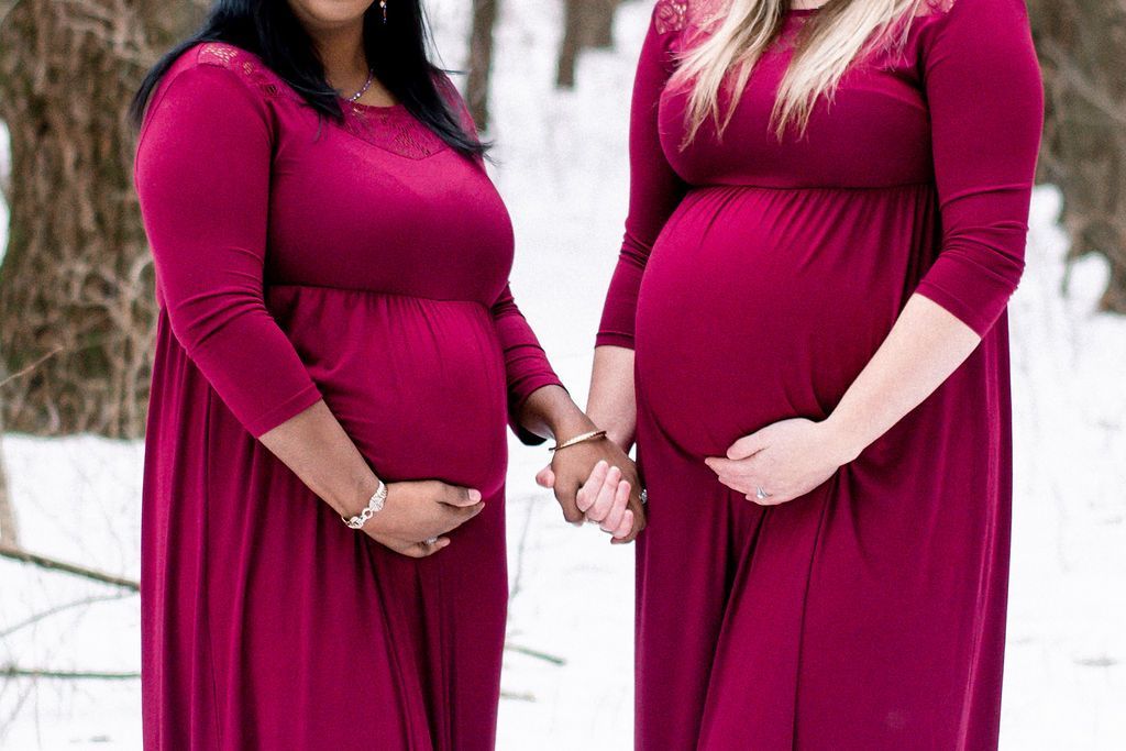 Two pregnant people in magenta dresses hold hands outdoors in a snowy setting.