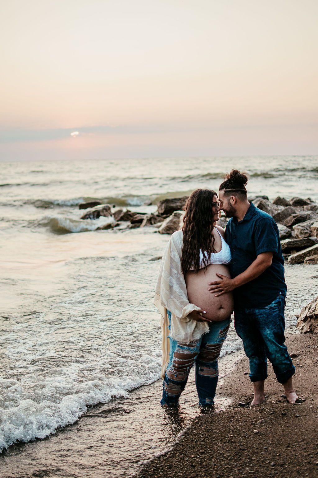 Couple standing on a rocky beach at sunset, embracing and facing each other by the waves.