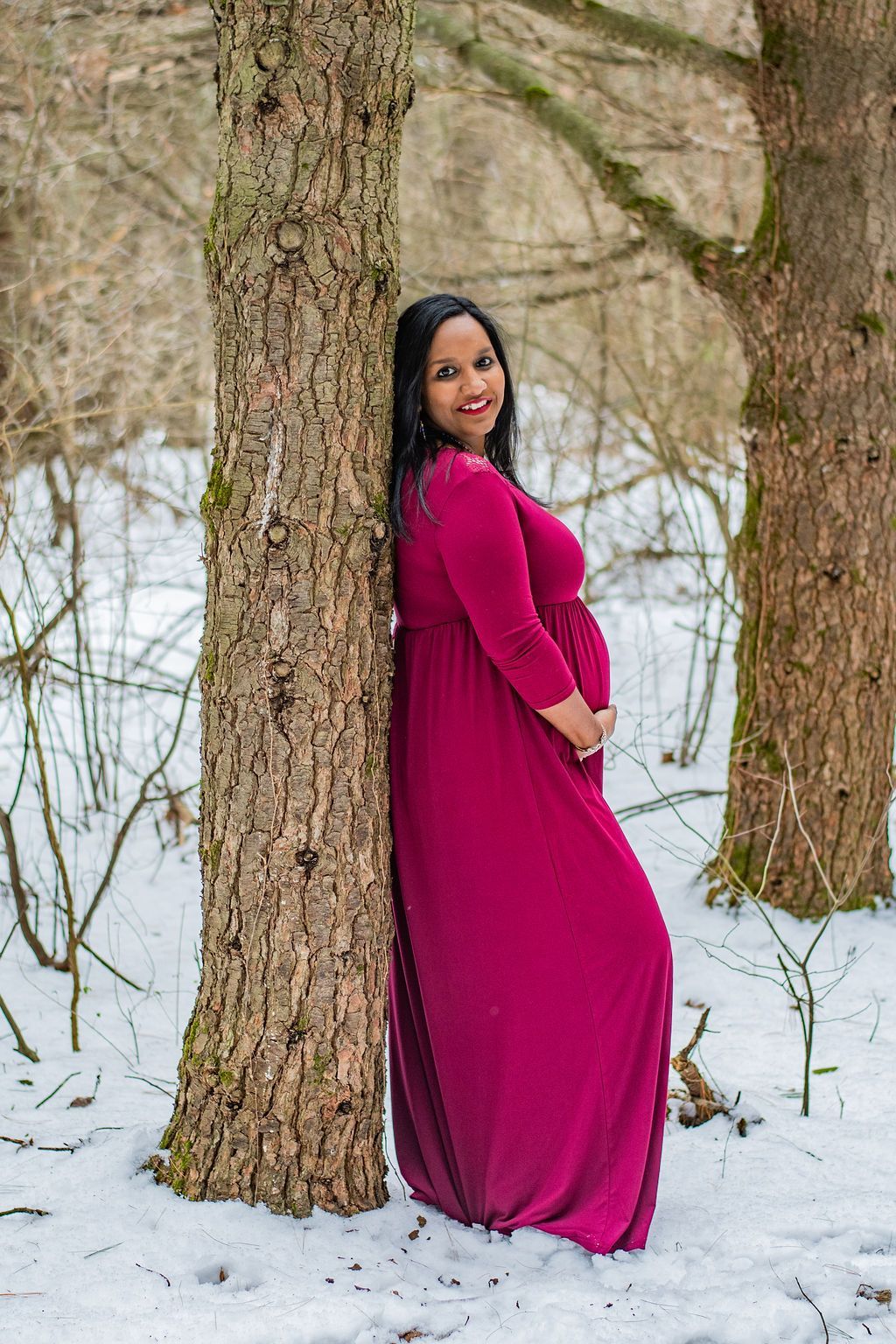 Woman in a magenta dress leaning against a tree in a snowy forest