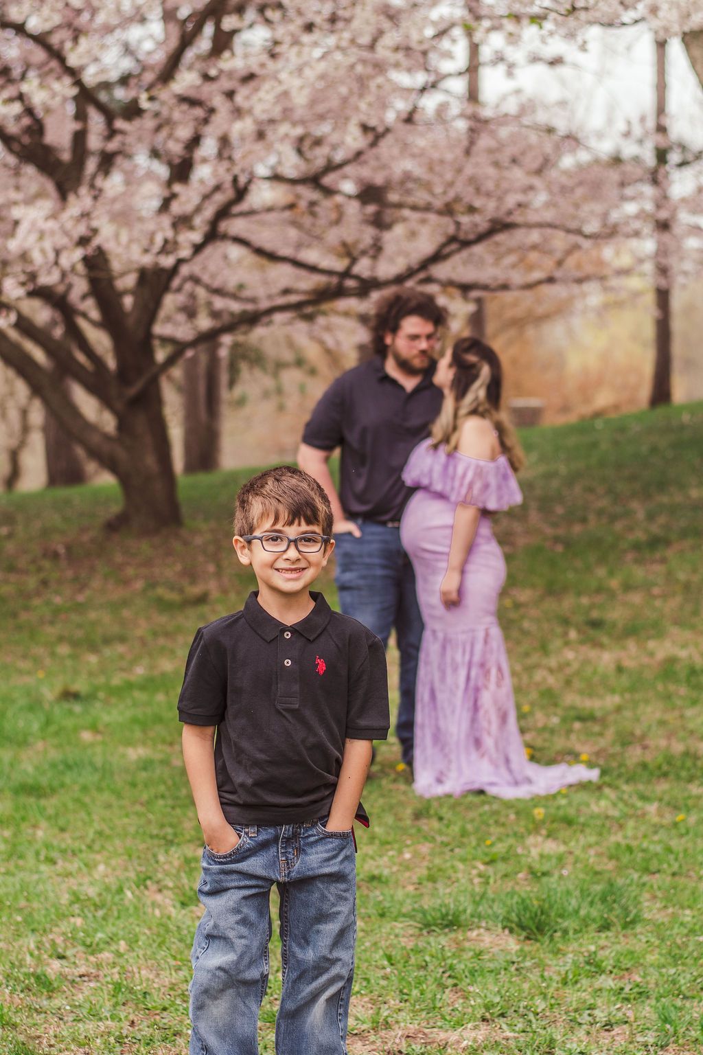Smiling child in glasses stands in front of two adults under blooming cherry trees in a park.