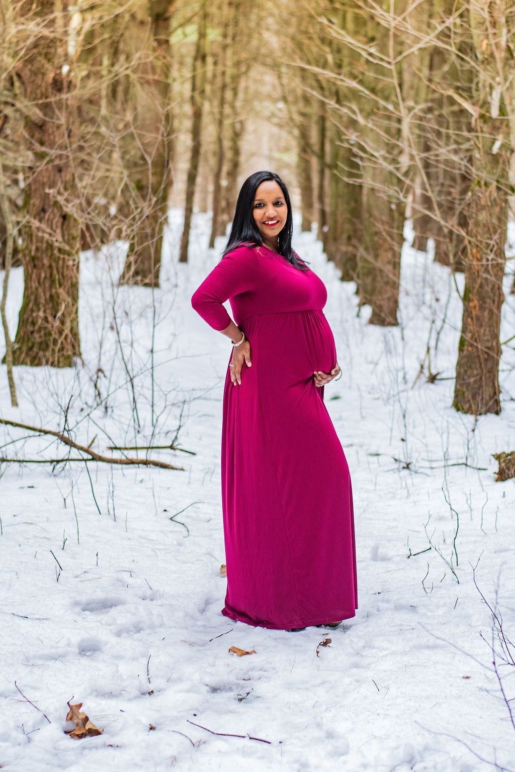 Woman in a bright magenta dress standing in a snowy forest path with bare trees