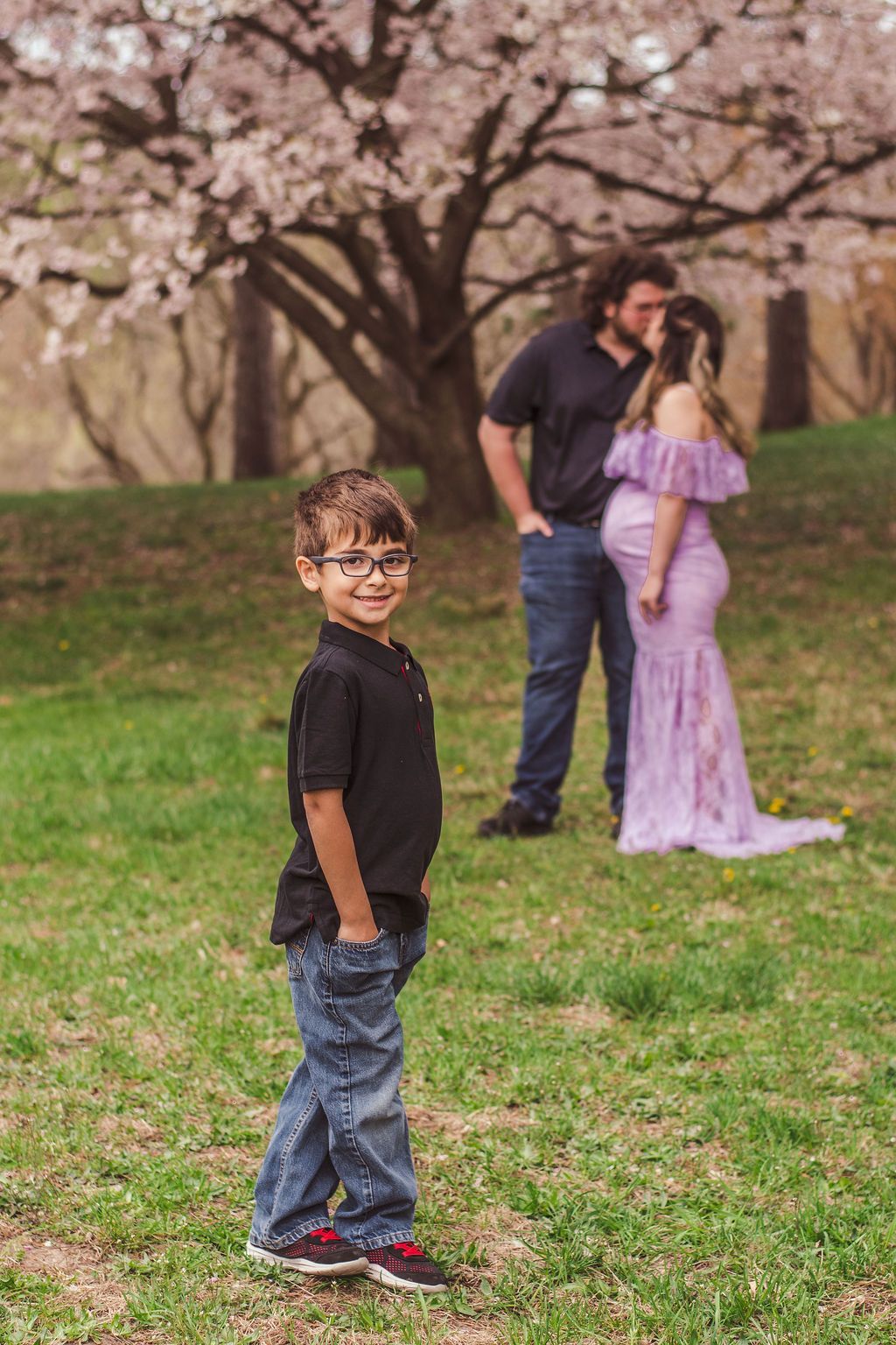 Young boy standing in a grassy orchard with blossoming trees and two people blurred in the background