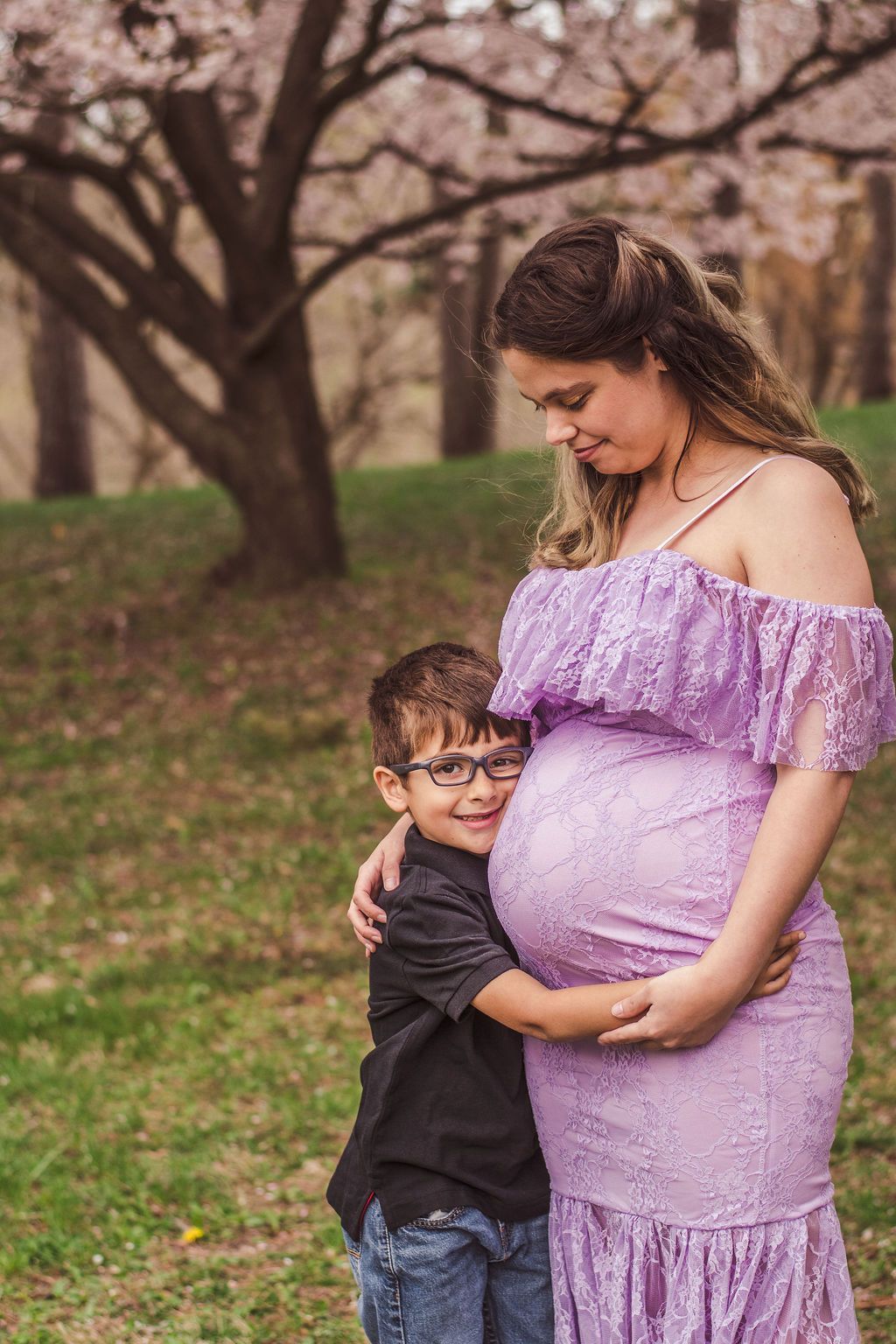 Pregnant woman in a lavender dress hugging a smiling child in a park with blooming trees