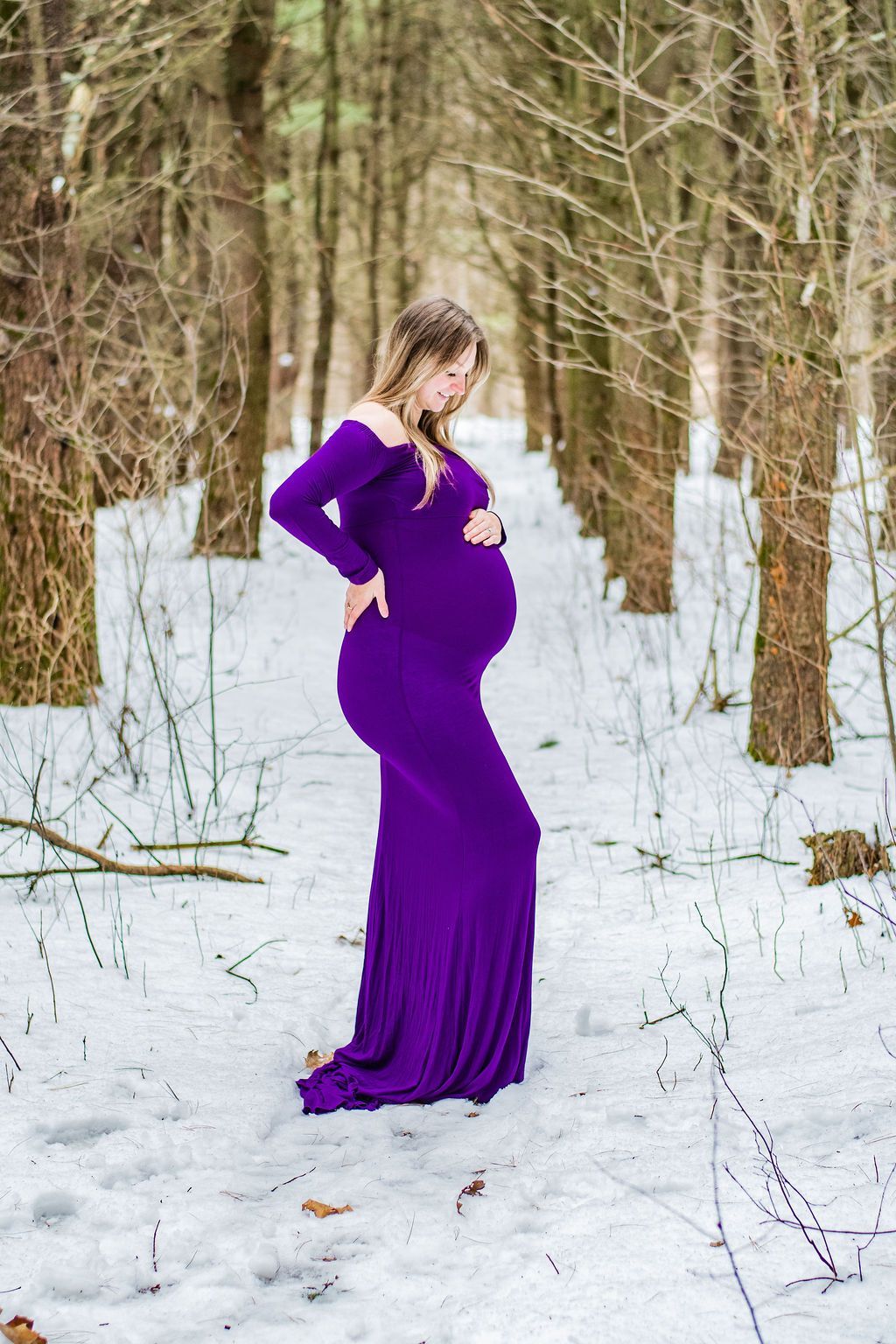 Pregnant person in a purple dress standing in a snowy forest