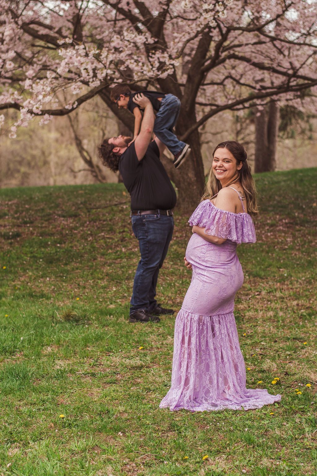 Pregnant woman in a lavender dress posing outdoors while a man lifts a child on his shoulders under blooming trees