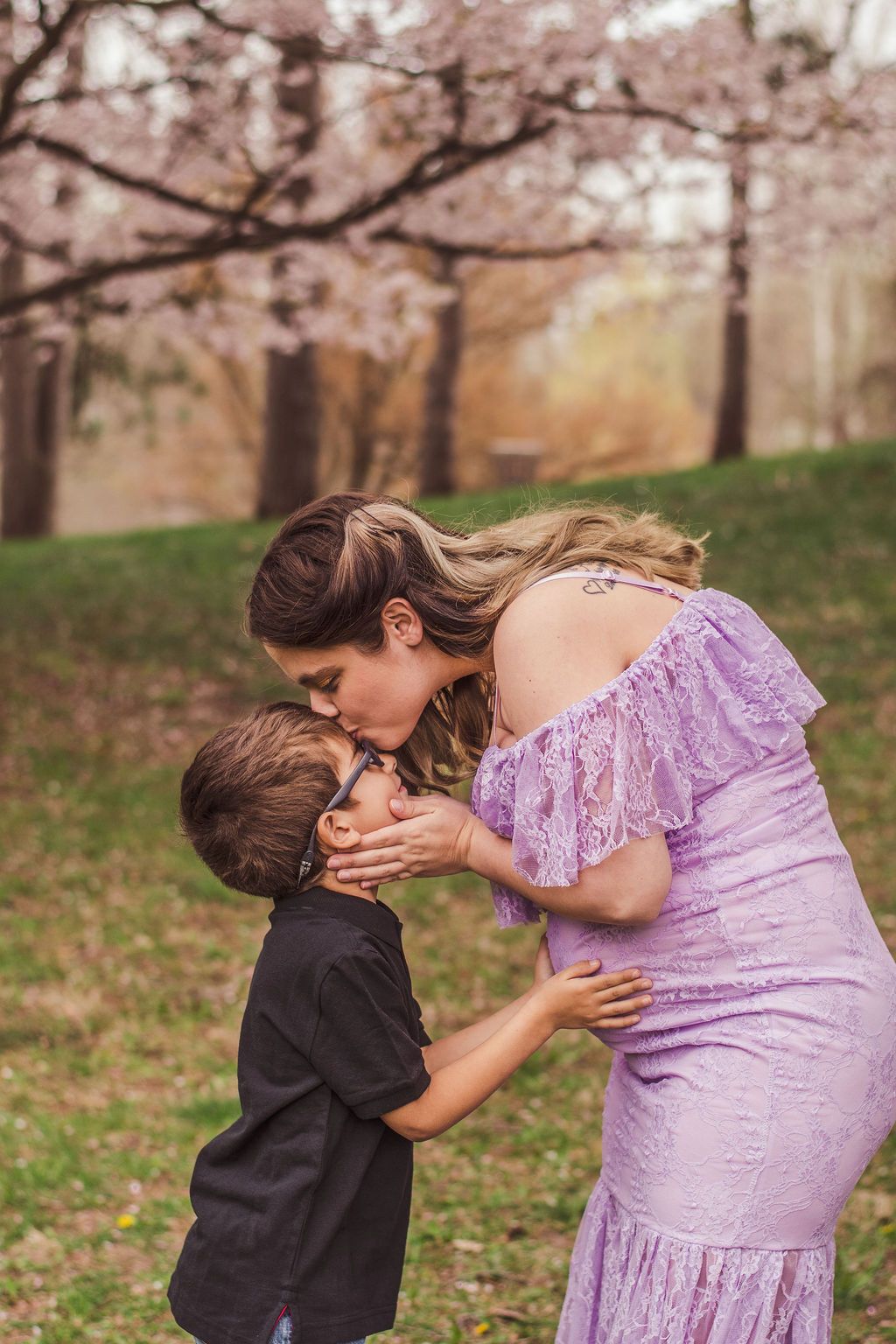 Woman in a lavender dress kisses a child outdoors under blooming trees.