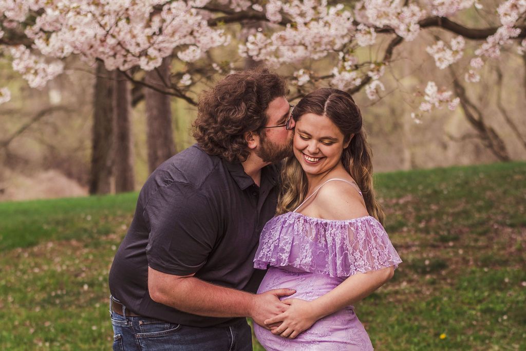 Couple sharing a kiss under blooming cherry trees, with the woman in a lavender dress smiling.