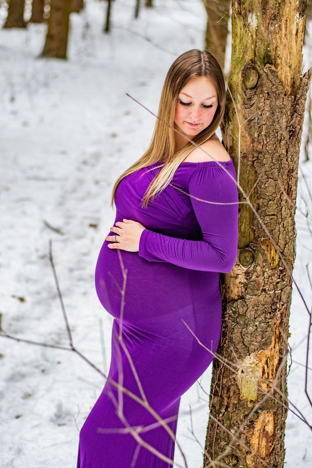 Pregnant woman in a purple dress leaning against a tree in a snowy outdoor setting
