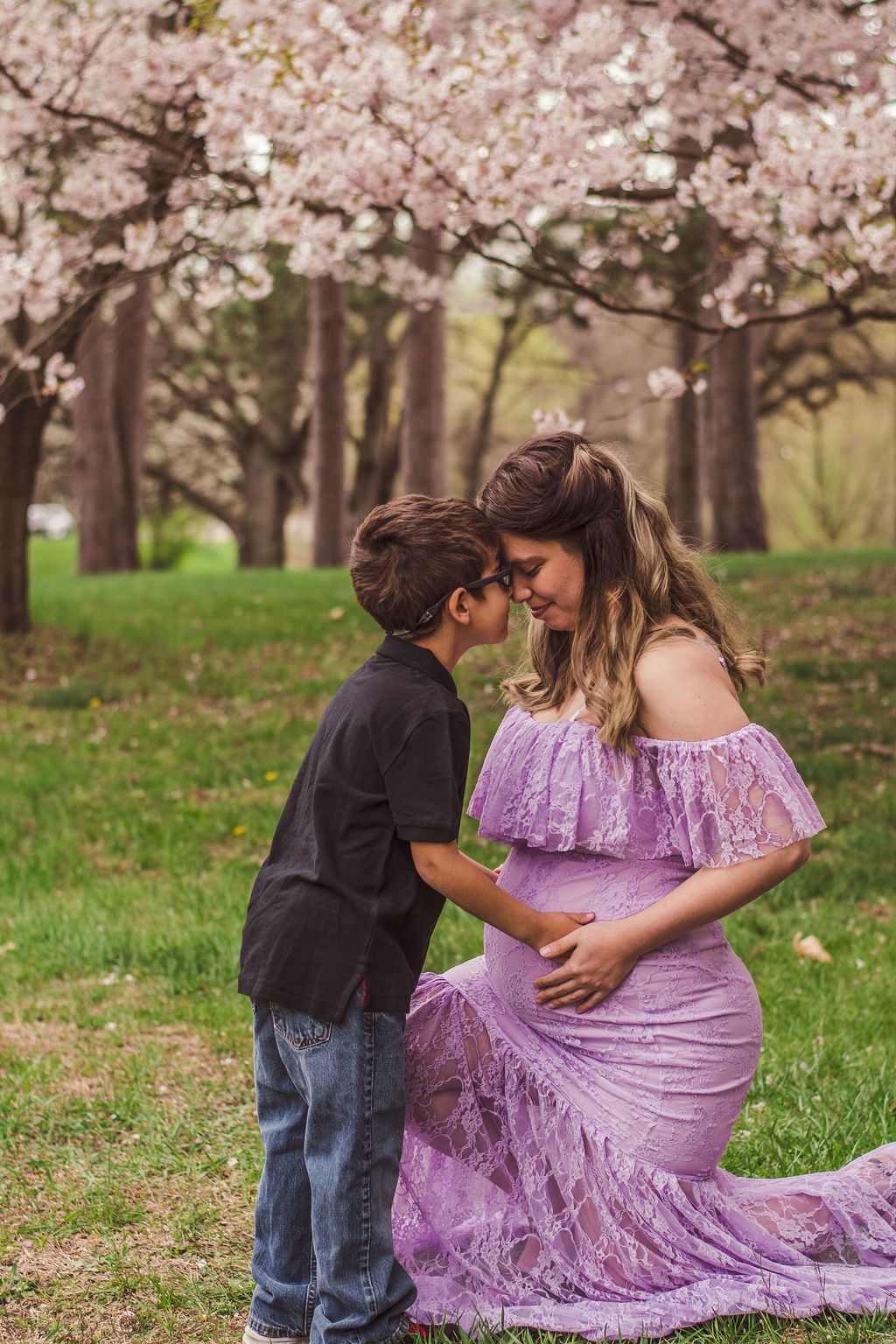 Woman kneeling in a lavender dress with a child in a black shirt, touching foreheads under blooming trees.