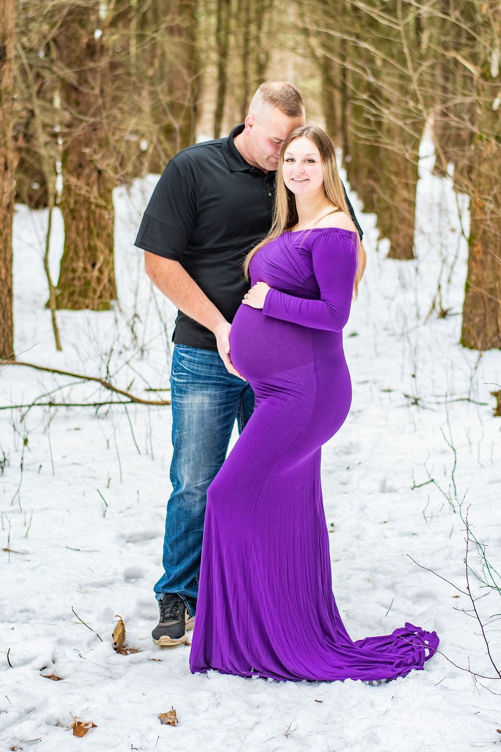 Couple posing outdoors in snow, with a woman in a purple dress and a man in jeans and a black shirt