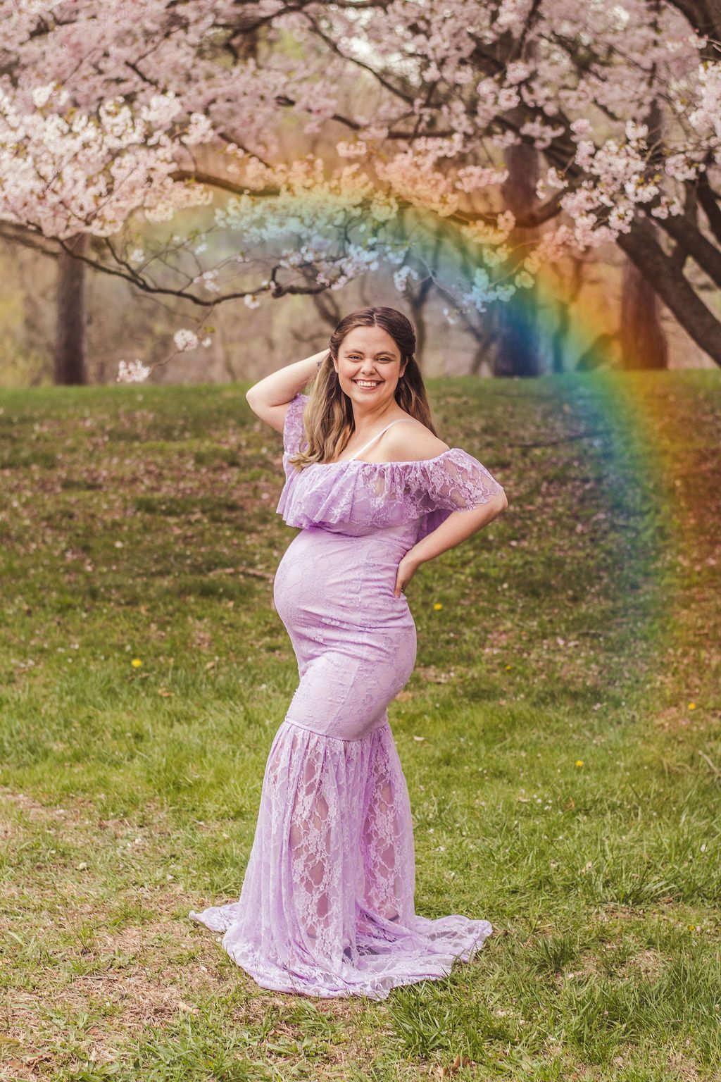 Pregnant woman in a lavender gown posing outdoors beneath blooming cherry blossoms with a rainbow backdrop