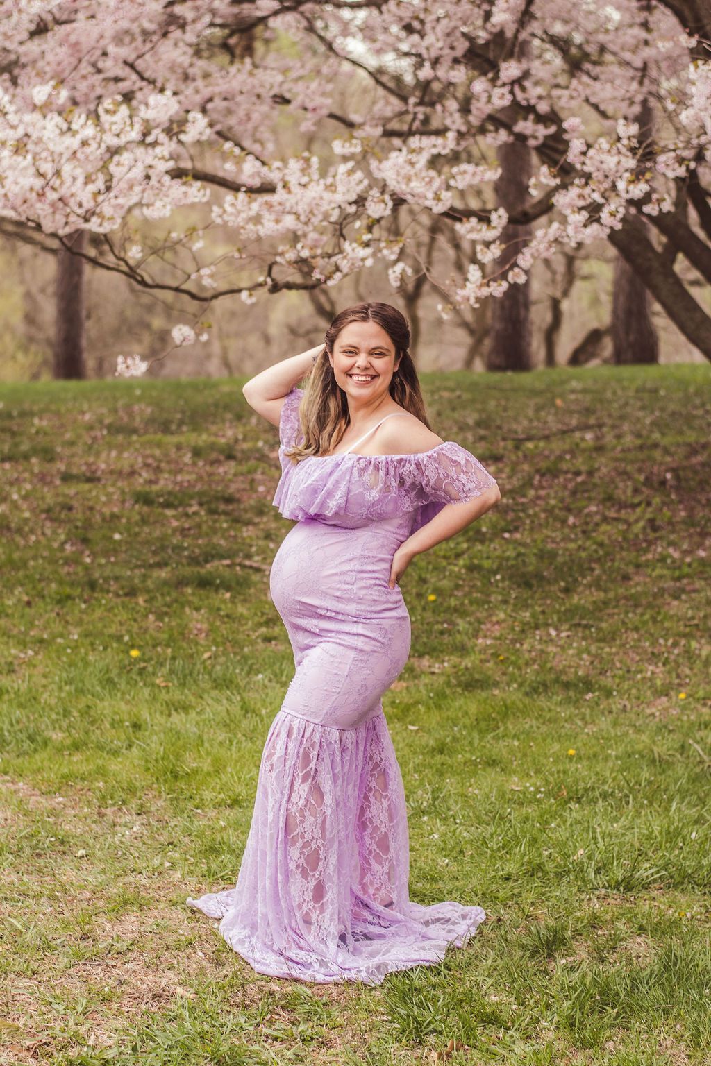 Pregnant woman in a lavender dress posing under blooming cherry trees in a grassy field