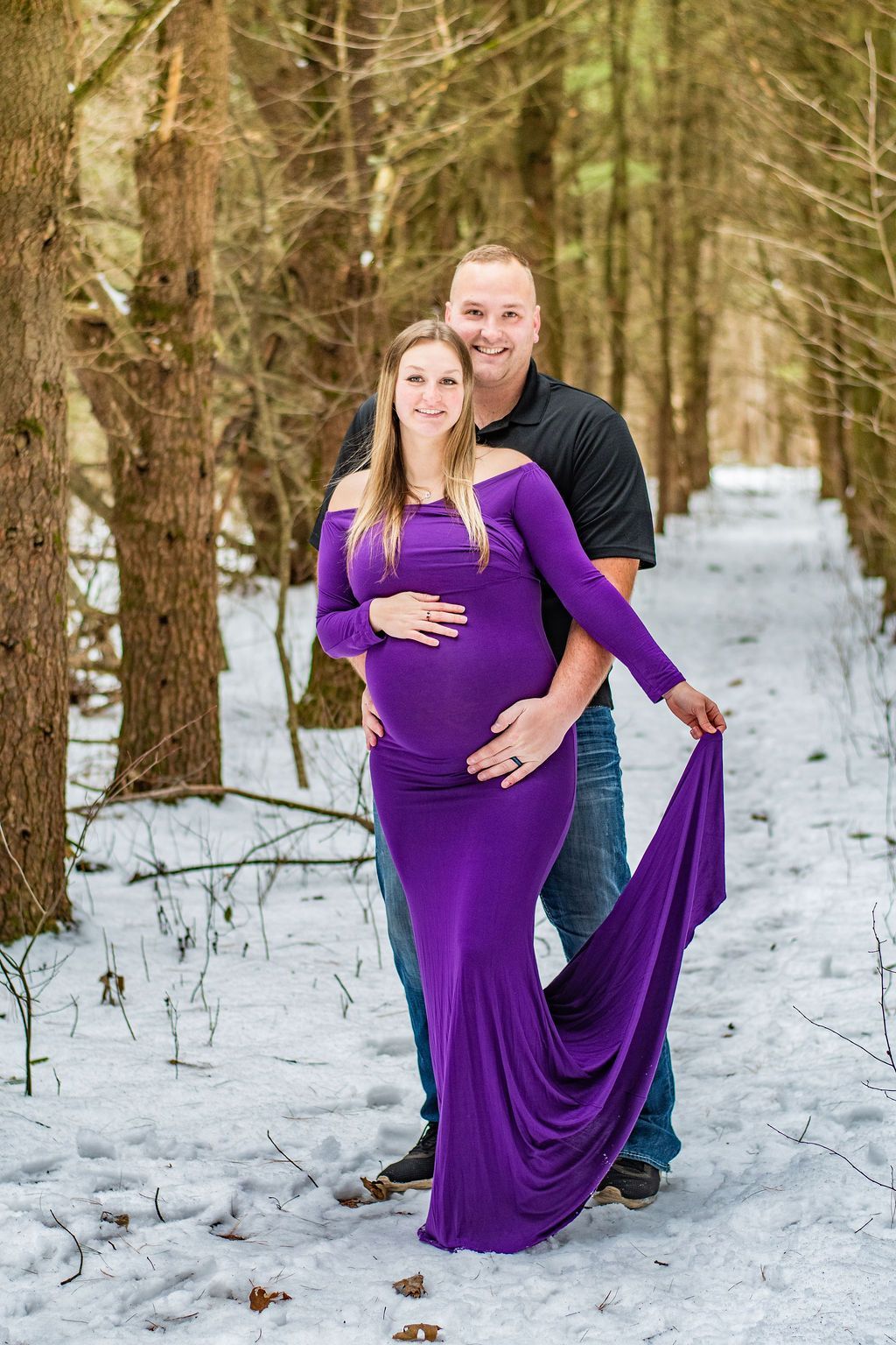 Couple standing on a snowy forest path, woman in a purple dress in front of man in black shirt