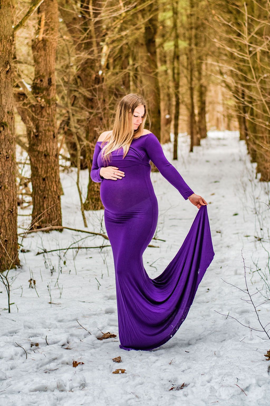 Pregnant woman in a purple gown standing in a snowy forest, holding the dress fabric