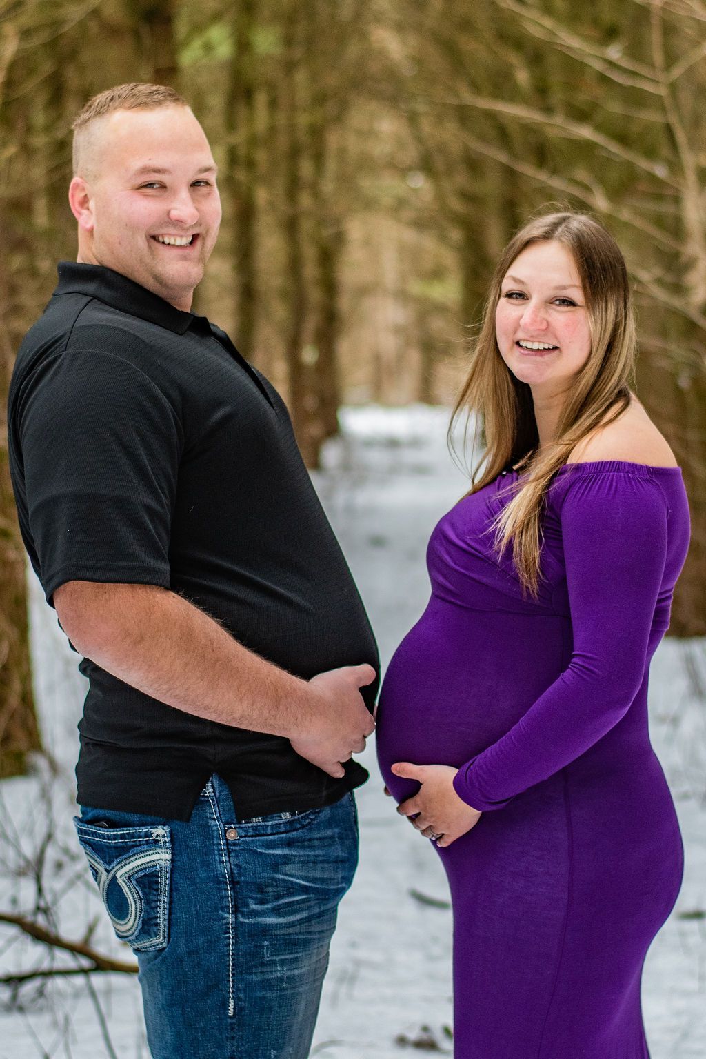 Pregnant couple standing in a snowy forest, smiling and holding the baby bump.