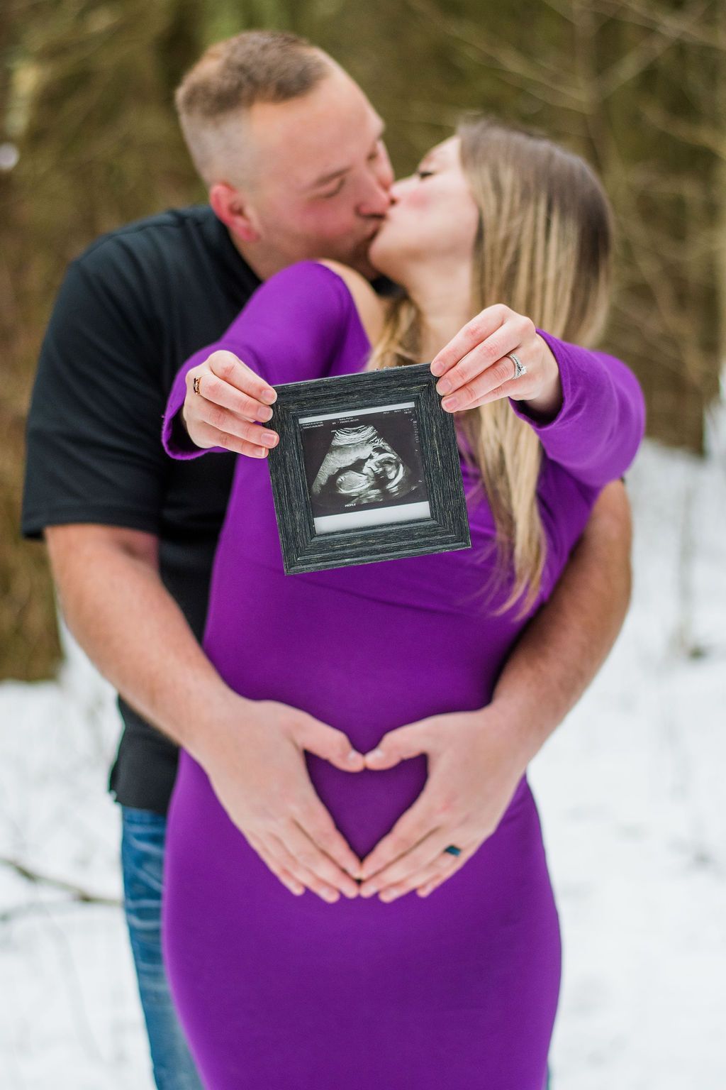 Couple embracing in snow, kissing while holding up an ultrasound photo in front of the pregnant partner.