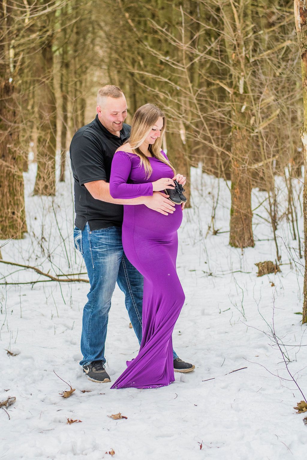 Couple standing in snowy woods, woman in purple dress holding a camera while man embraces her from behind