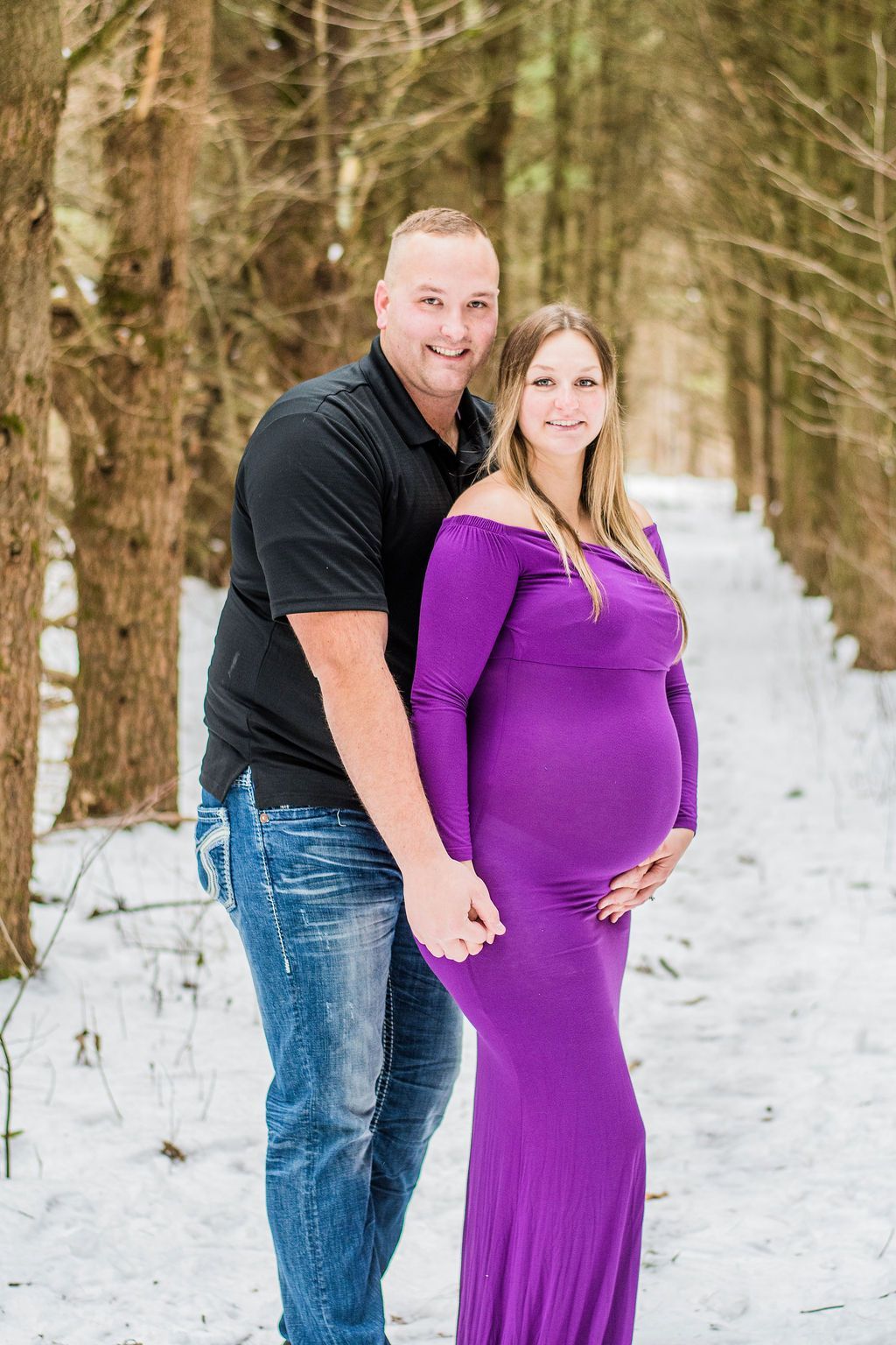 Couple standing on a snowy path, smiling and posing; woman in purple dress with hands on her belly.