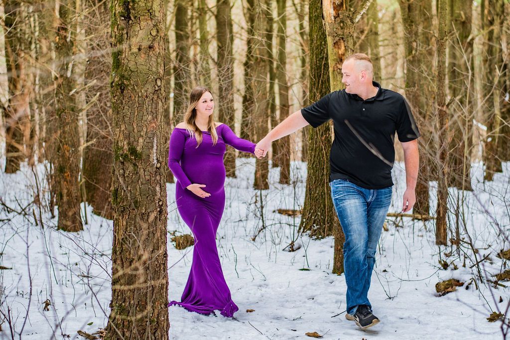 Couple holding hands in a snowy forest, with the woman in a purple dress and the man in a black shirt and jeans