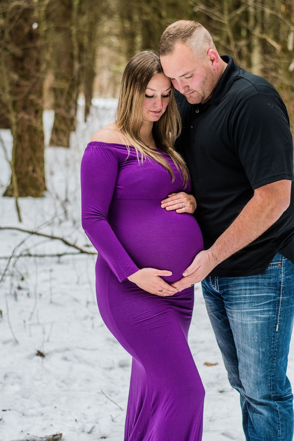 Pregnant couple standing in snowy woods, with the man embracing the woman's belly.