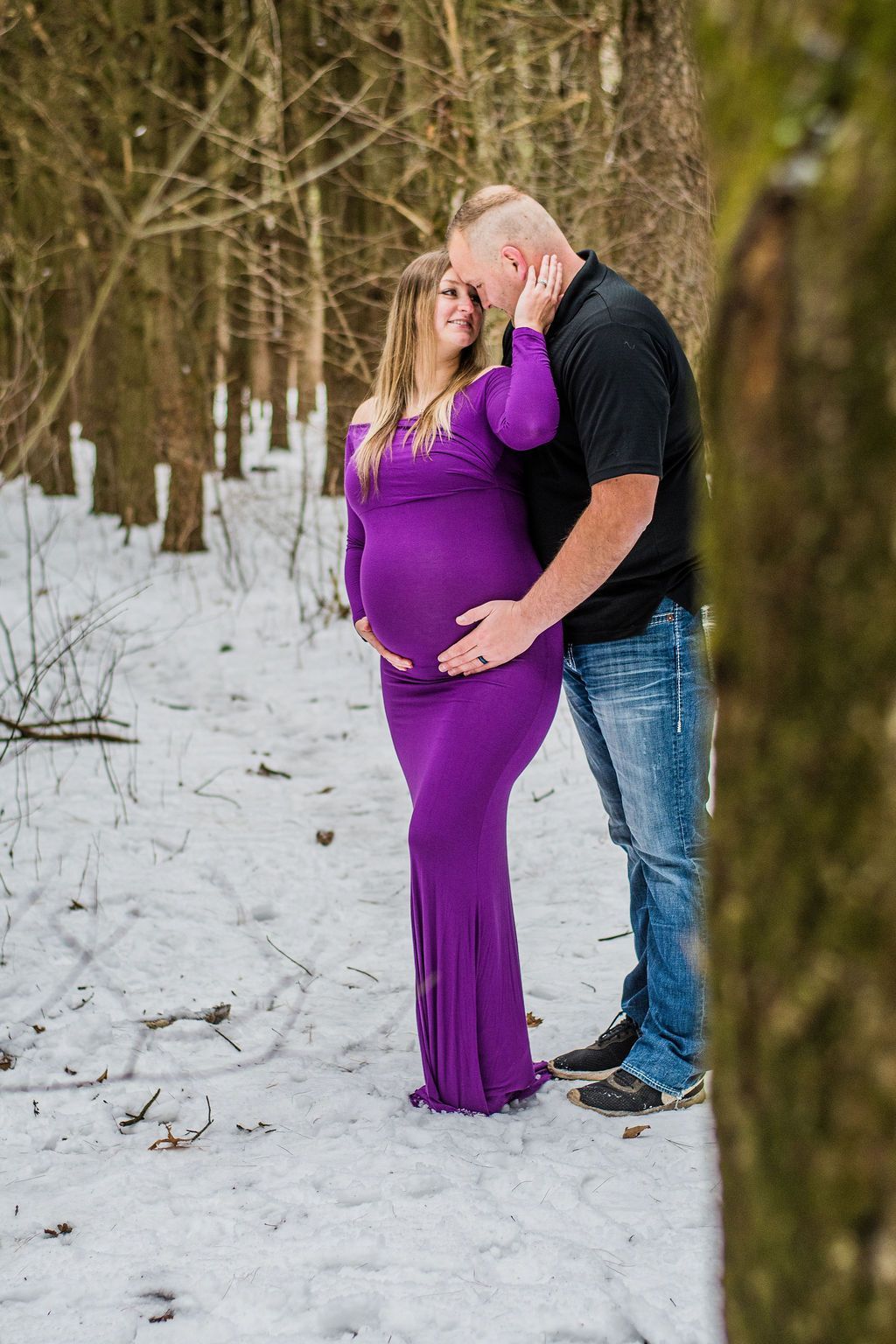 Couple embracing outdoors in snowy woods, woman in purple dress and man in black shirt and jeans