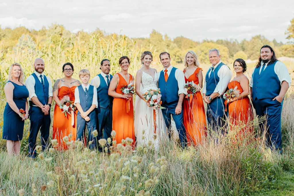 Group of people in bright formal outfits standing in a grassy field with autumn trees behind them