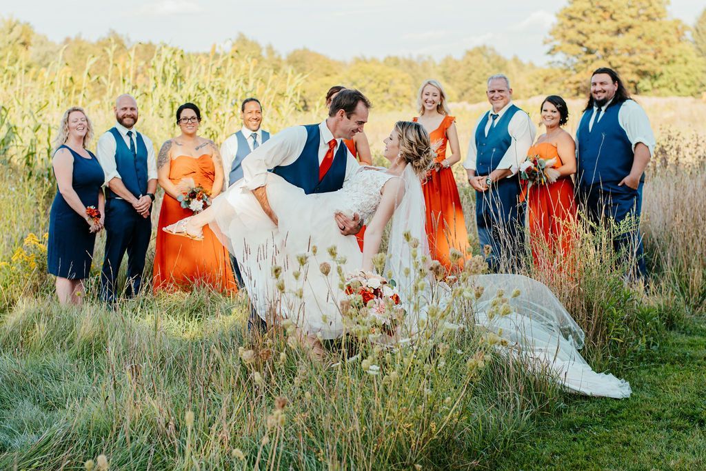 Wedding party in colorful attire posing in a grassy field, with the couple kissing in front.