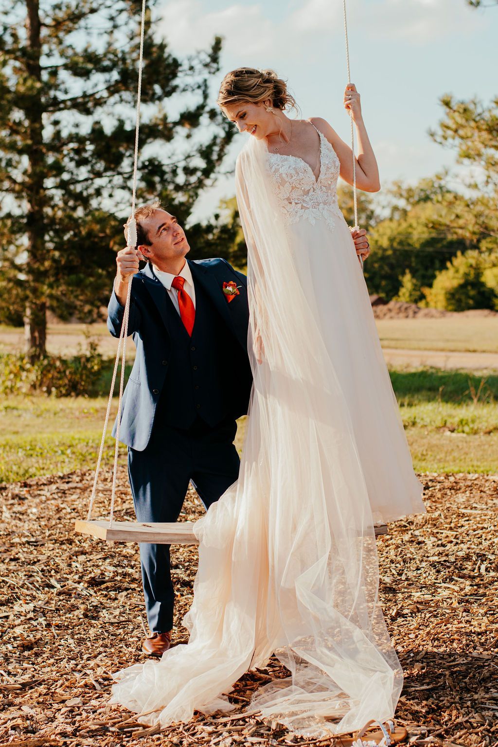 Bride in a flowing white dress and groom in a dark suit swing together outdoors on a sunny day