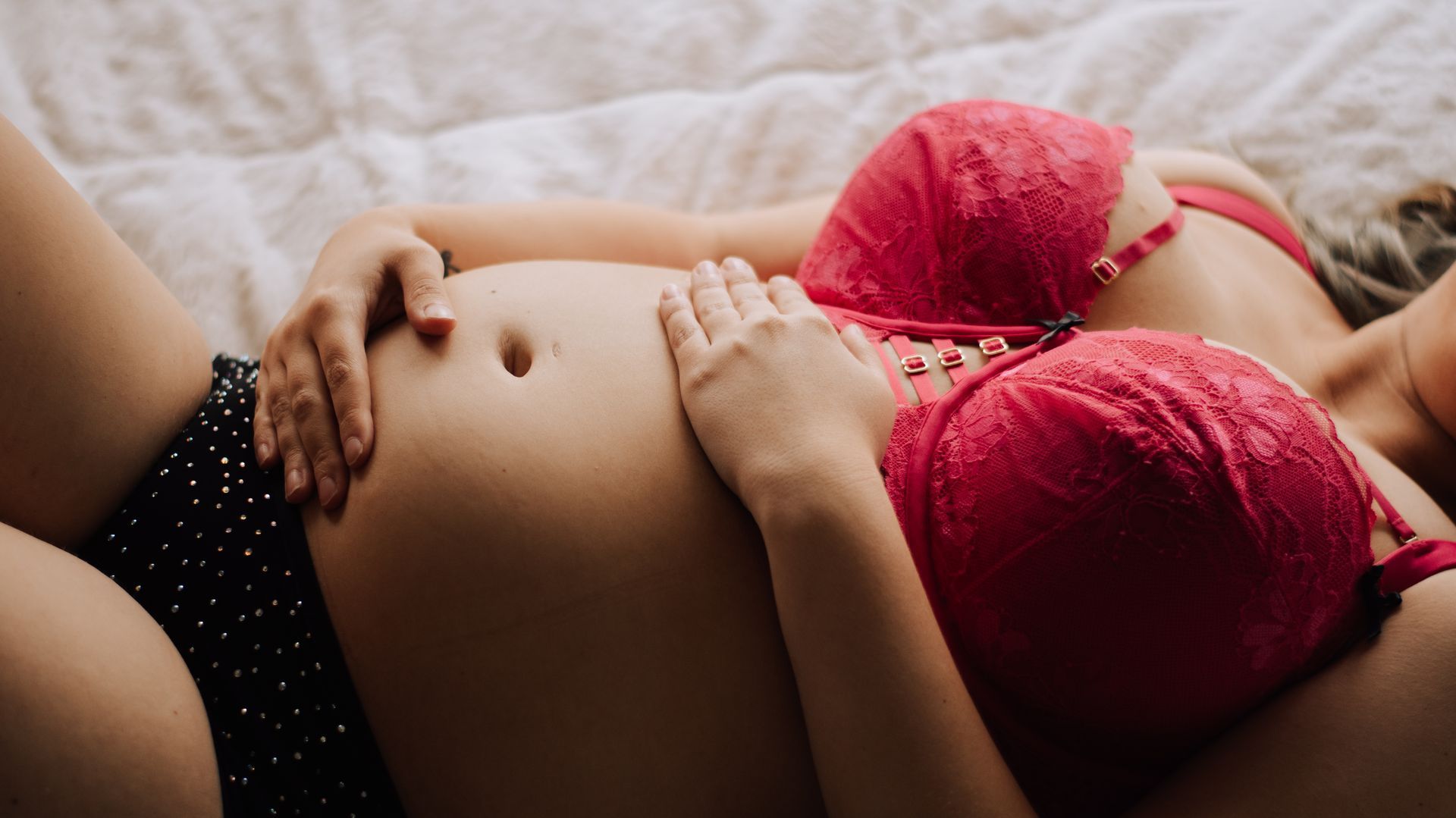 Pregnant person lying on a bed, wearing a red bra and black underwear, hands resting on belly