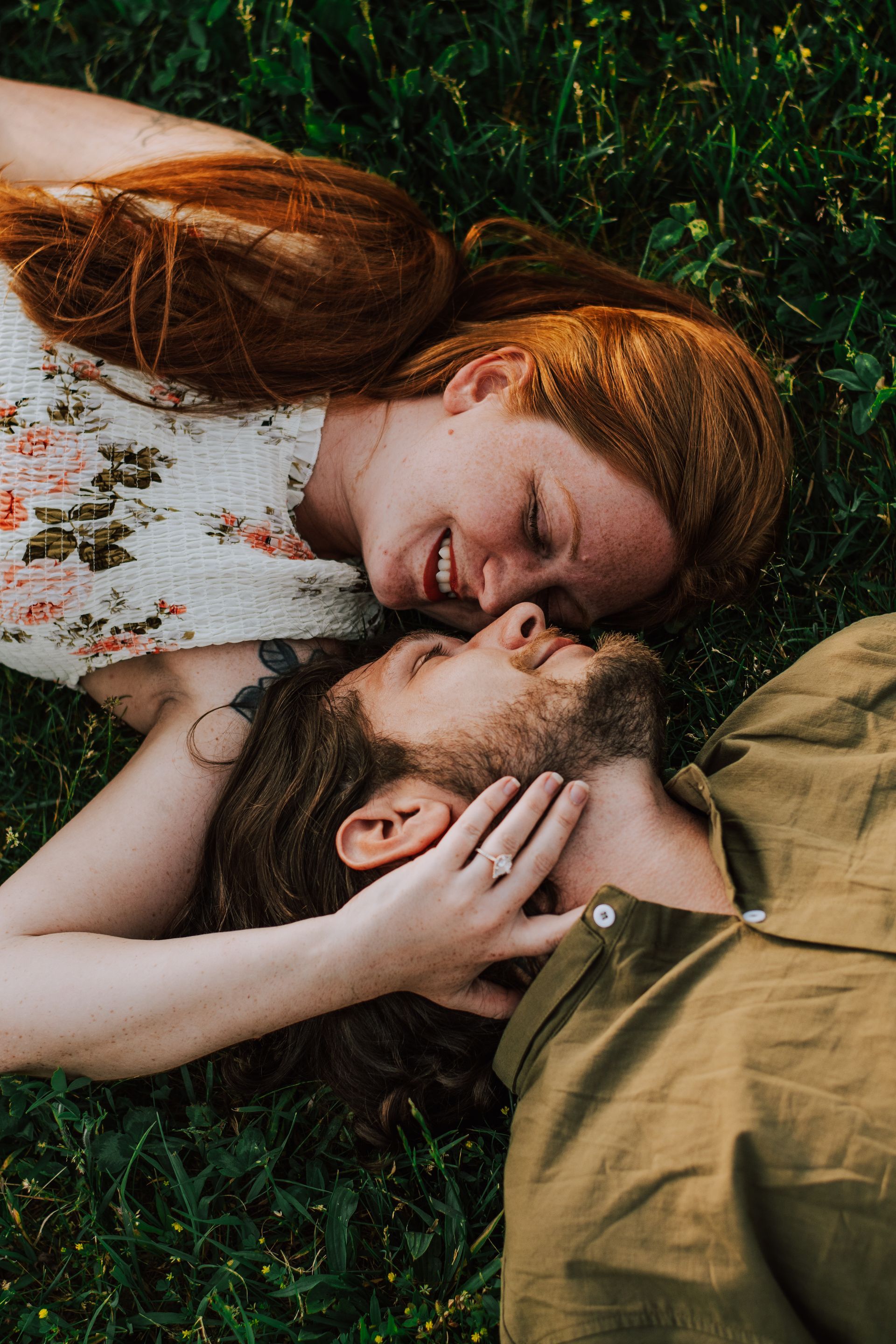 Couple lying on grass, forehead to forehead, smiling softly in a warm outdoor embrace.