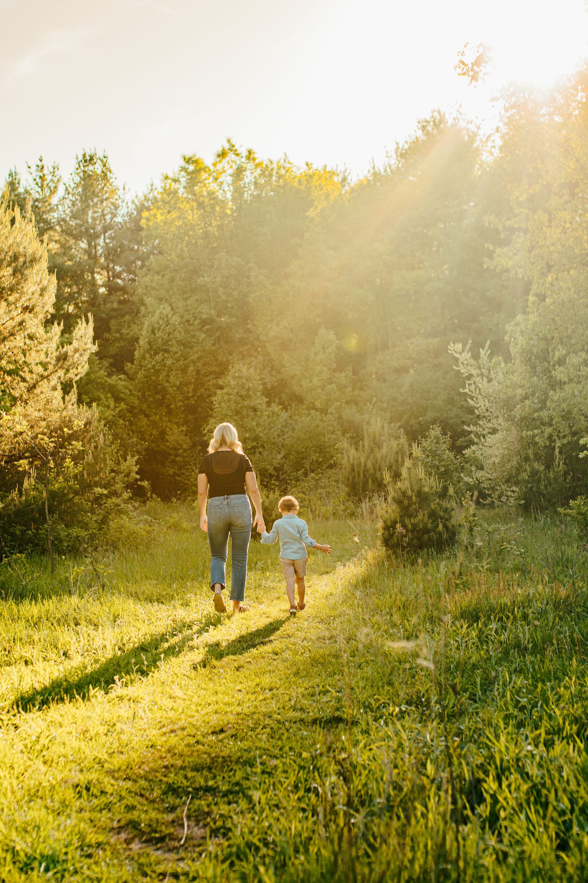 Two people walking hand in hand through a sunlit grassy path surrounded by trees