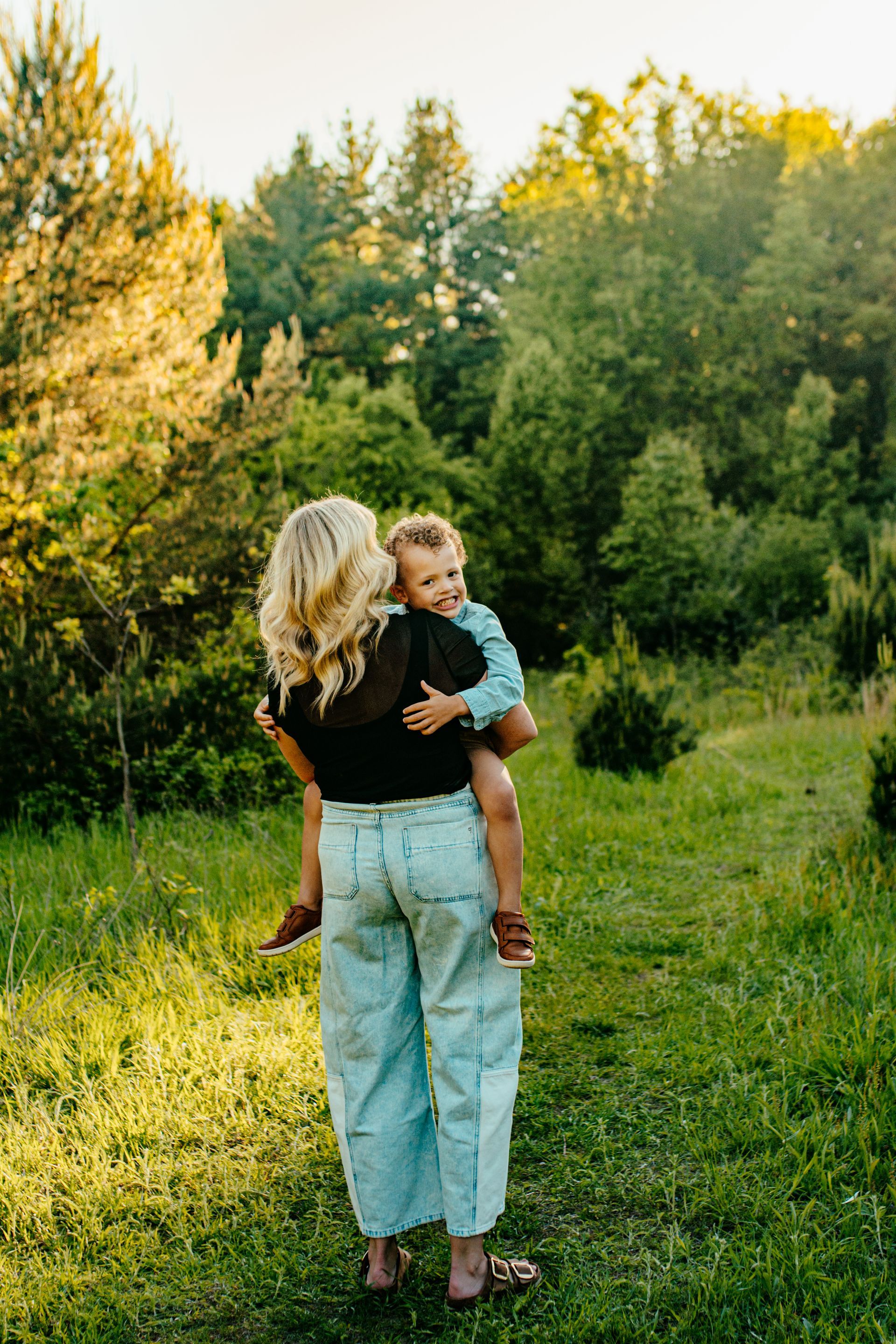 Woman holding a small child in a grassy field at sunset with trees in the background