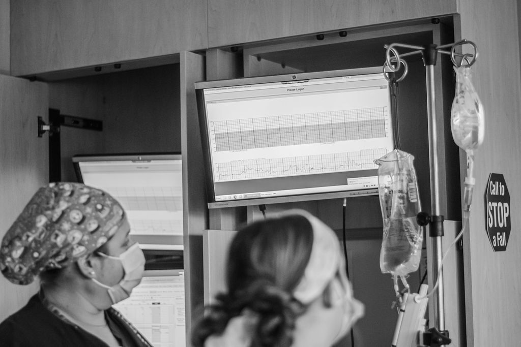Two masked healthcare workers look at an IV drip and monitor beside a patient bed.