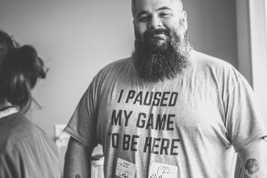 Bearded man in a T-shirt reading “I PAUSED MY GAME TO BE HERE,” standing beside a woman indoors