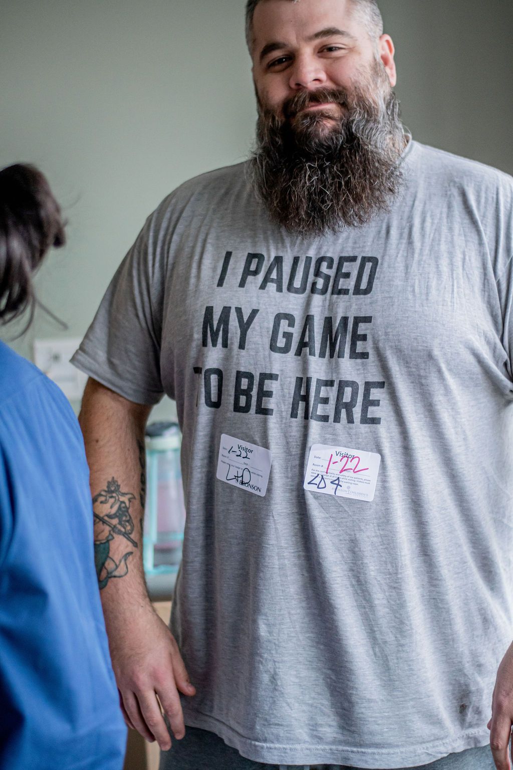 Man in gray T-shirt reading “I paused my game to be here,” standing indoors beside another person