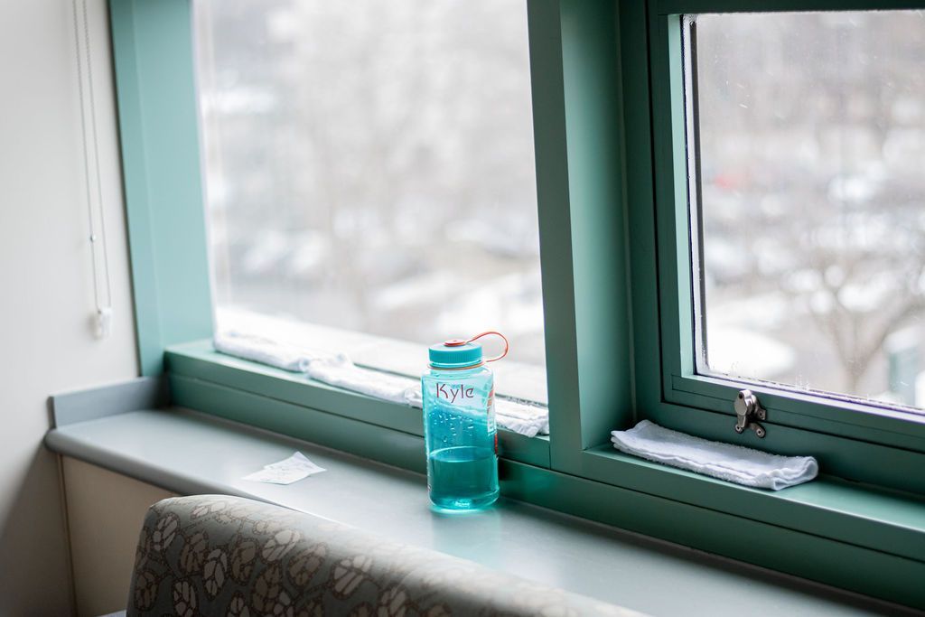 A teal jar sits on a windowsill beside green-framed windows and a patterned cushion.