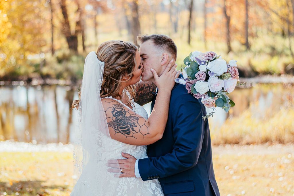 Bride and groom kissing outdoors beside a lake, with autumn trees and a bouquet in the background