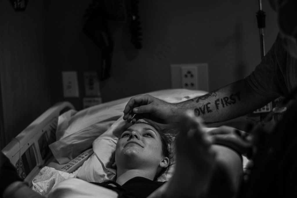 Hospital patient lying in bed while a caregiver holds their forehead in a dim room